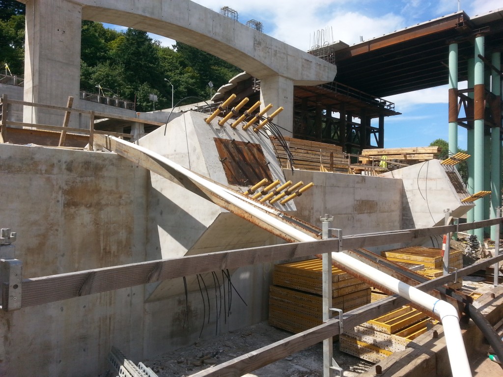 This is the support structure where the steel arch will touch down on the west bank.  Those black cable-looking things next to the yellow rods aren’t structural, but they are vital.  As concrete sets after pouring, it heats up.  In a block of concrete as big as this, the heat in the center of the block can’t make it to the surface to be released.  If the heat can’t dissipate, the concrete will be weaker when it eventually sets.  Those “cables” are actually tubes to carry water through the concrete, cooling the core and allowing uniform setting.